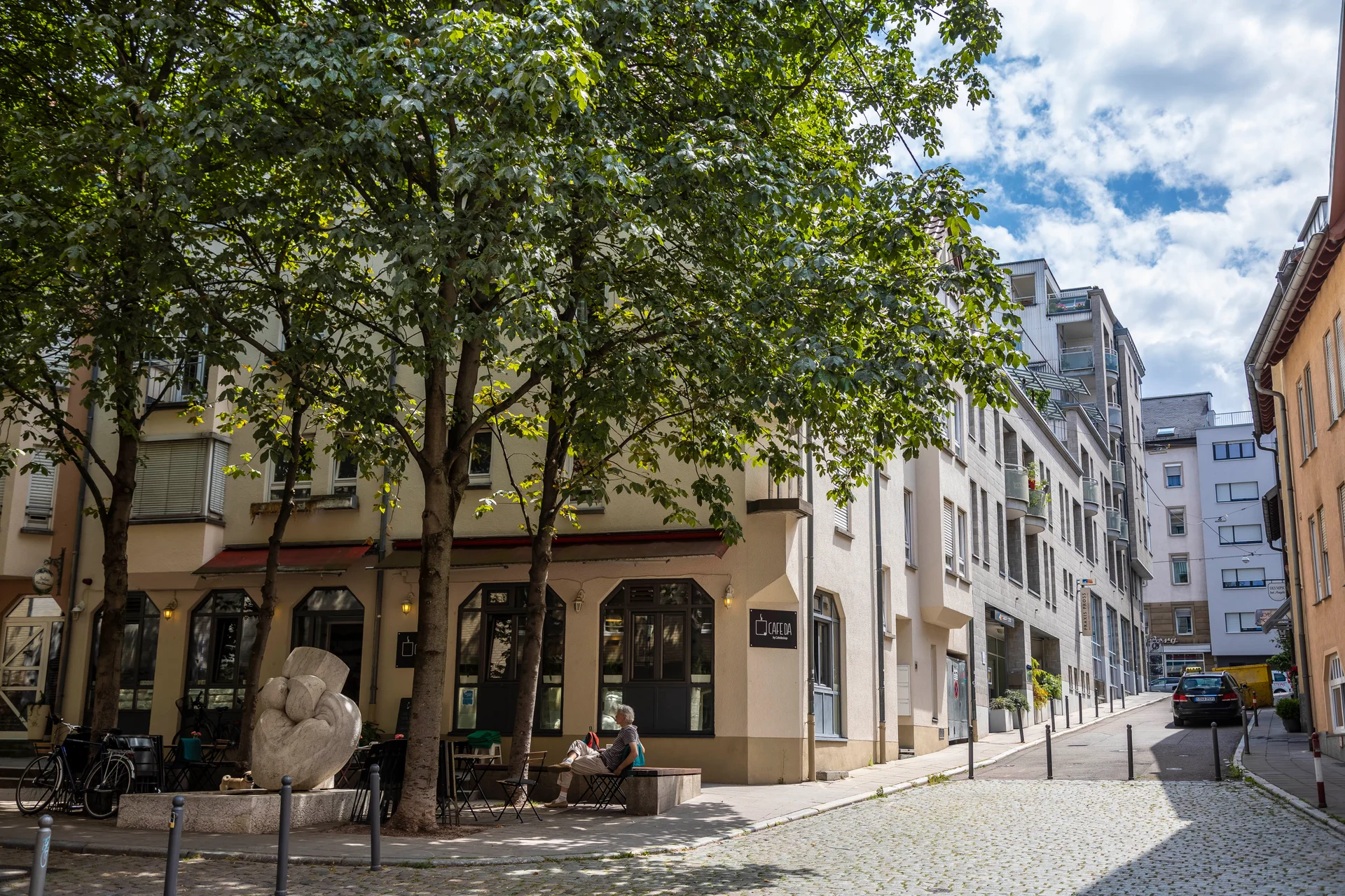 Straßenszene mit Bäumen, Sitzbank, Skulptur und Personen vor einem Eckgebäude in einer ruhigen Gasse.