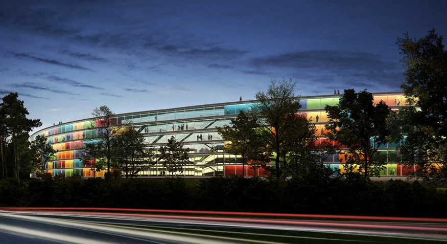 Das Eiermann Campus Schleifenhaus mit einer geschwungenen Glasfassade, die in mehreren Farben beleuchtet ist, vor einem Abendhimmel und Bäumen im Vordergrund.