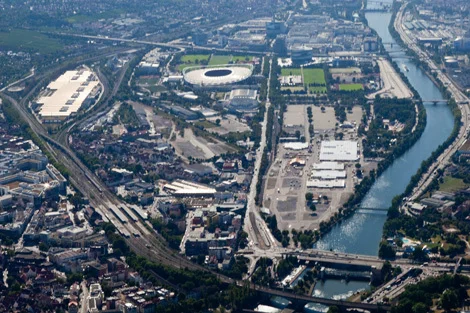 Luftaufnahme der Fläche des entstehenden Stadtquartiers Neckarpark mit Fluss, Brücken, Stadion und umliegenden Gebäuden und Straßen.